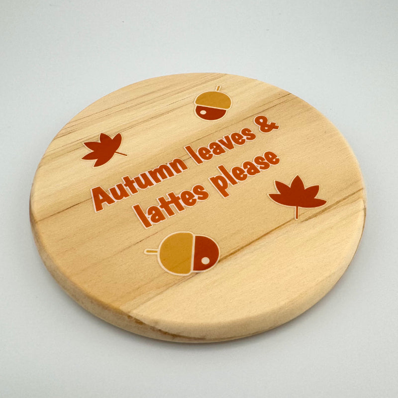 Wooden coaster with 'Autumn leaves & lattes please' text and leaf designs on a light gray background